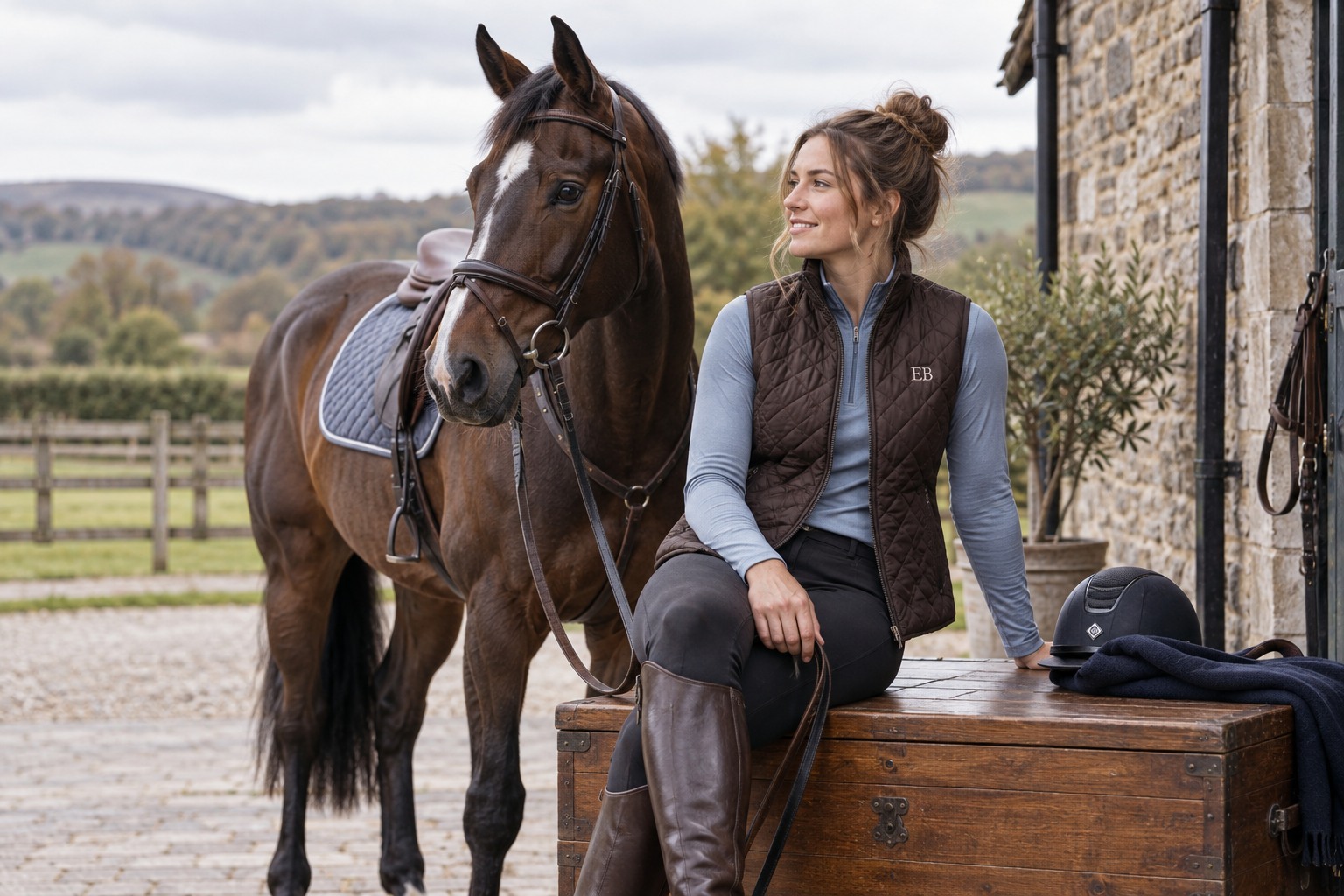 An adult rider in a dark brown quilted gilet and a soft blue base layer sitting casually on a solid wooden tack trunk.